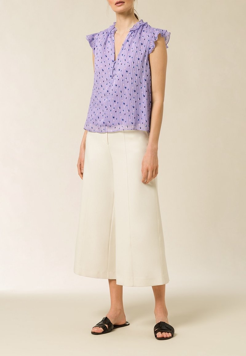 Blusa lavanda con maniche a volant, tessuto testurizzato e motivi a cuore blu, abbinata a pantaloni bianchi a gamba larga e sandali neri.