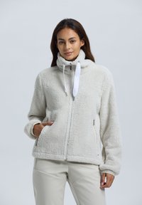 Nainen, jolla on päällään vaalea harmaa fleece-tyyppinen takki, jossa on korkea kaulus ja yksi käsi taskussa, seisoo tasaista taustaa vasten.