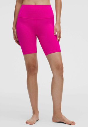 Mujer con shorts de ciclista de talle alto de color rosa brillante de pie descalza frente a un fondo claro y liso.