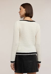 Donna di spalle che indossa un maglione bianco peloso con bordi neri e una mini gonna di pelle nera, con capelli castani diritti e corti e orecchini d'oro.