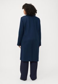 Una persona con capelli ricci si trova con le spalle alla fotocamera, indossando un lungo cappotto blu navy e pantaloni blu navy larghi su uno sfondo semplice.