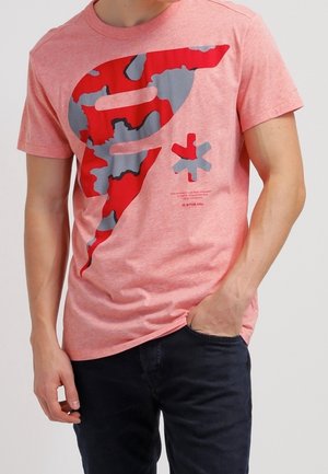 Homme portant un t-shirt rose à manches courtes avec un grand motif abstrait rouge et gris et un pantalon noir, main dans la poche.