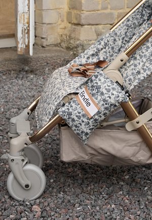 Passeggino grigio chiaro con tessuto floreale, cinghie color cuoio e borsetta floreale coordinata con etichetta "Elodie" su superficie ghiaiosa.