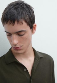Polo verde scuro con colletto e bottoni. Il tessuto appare morbido con una texture liscia. Il modello ha capelli corti e texturizzati.
