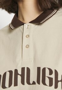 Beige poloskjorte med brun krage som har to striper, tre klare knapper og mørkebrun tekst på framsiden. Glatt bomullstekstur.