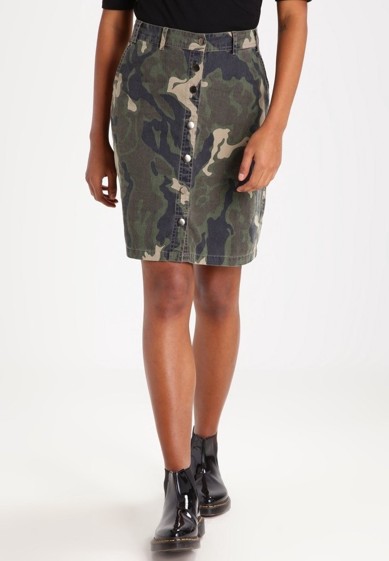 Falda de mezclilla de camuflaje con forma ajustada, frente con botones y un dobladillo sin terminar. Combinada con botas negras brillantes hasta el tobillo.