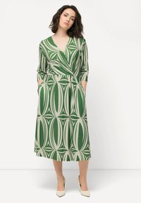 Grünes Kleid mit einem geometrischen Muster in Creme, mit tiefem V-Ausschnitt, ¾-Ärmeln und Taschen. Glattes Material und knielanger Schnitt.