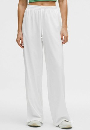 Persoon draagt losse witte broek met trekkoord en witte sneakers, staat tegen een effen lichte achtergrond.