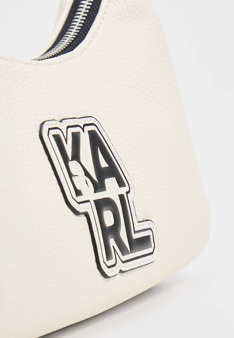Sac texturé blanc avec un patch noir et blanc "KARL" et une fermeture éclair en argent visible sur le bord supérieur, le tout sur fond blanc.