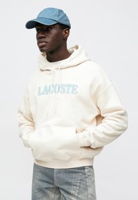 Hoodie creme com o texto "LACOSTE" em azul, com bolso canguru e cordões, combinado com um boné de ganga claro e calças estampadas.