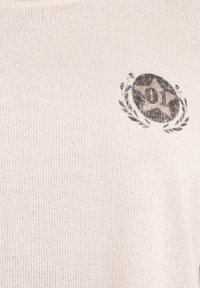 Suéter de punto color beige claro con cuello redondo y un logo circular negro con diseño de estrella y laureles en la parte superior izquierda.