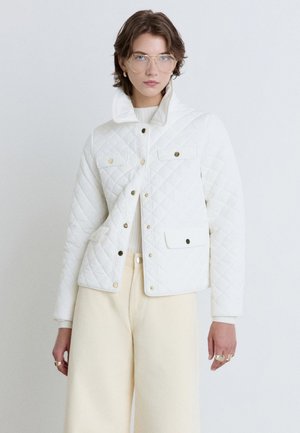 Person trägt klare Brille, eine weiße gesteppte Jacke mit goldenen Knöpfen, cremefarbene Hose und goldene Ringe, steht vor einem schlichten hellen Hintergrund.