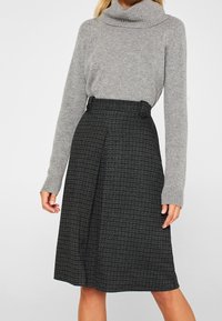 Pull gris à col bénitier avec des manches longues, associé à une jupe noire mi-longue à motif tartan et détail de boutons sur le côté.