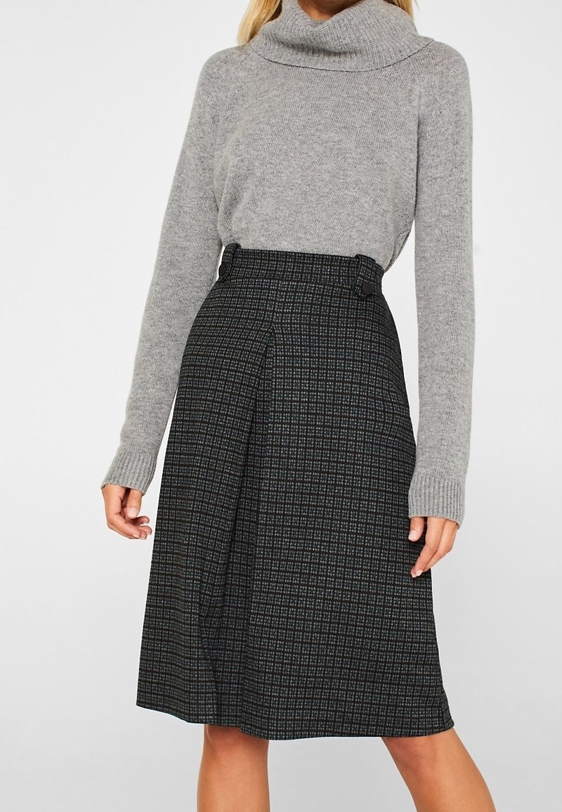 Pull gris à col bénitier avec des manches longues, associé à une jupe noire mi-longue à motif tartan et détail de boutons sur le côté.