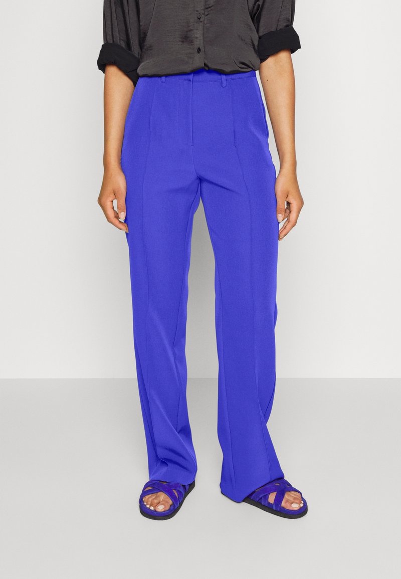 Personne portant un pantalon bien ajusté de couleur bleu vif, une chemise noire à manches retroussées et des sandales assorties bleues avec des brides croisées.
