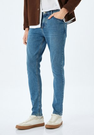 Jeans de mezclilla azul claro con un ajuste slim, diseño de cinco bolsillos y un ligero efecto de desgaste. Combinados con zapatillas en color crema y una chaqueta marrón con capas.