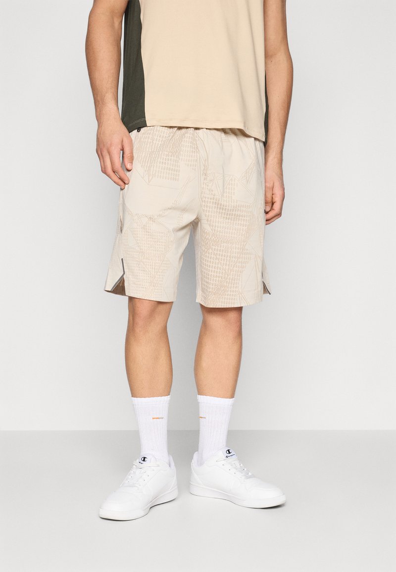Pantalones cortos beige con patrón, cintura elástica, bolsillos laterales y diseños angulares. Combinados con zapatillas blancas y calcetines de longitud media.