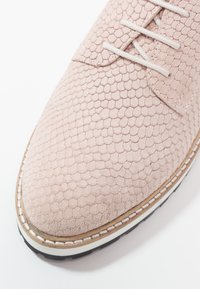 Chaussure à lacets rose clair avec motif hexagonal texturé, couture blanche le long de la semelle beige et semelle extérieure en caoutchouc noir.