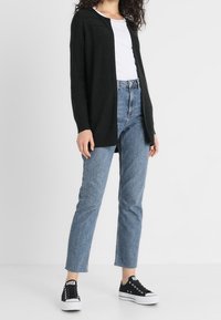 Kvinna iklädd en svart öppen cardigan, vit t-shirt, blå högmidjade jeans och svarta låga sneakers stående på ett vitt golv.