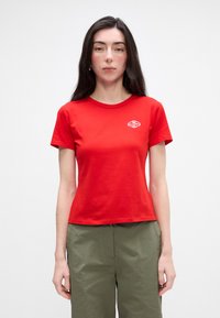 Crvena pamučna majica s okruglim izrezom, kratkim rukavima i bijelim izvezenim logoom na lijevoj strani prsa. Prilagođen dizajn, glatka tekstura.