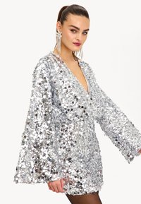 Vestido mini de lentejuelas plateadas con un escote en V profundo, mangas anchas y un corte relajado. Presenta lentejuelas circulares brillantes que cubren toda la superficie.