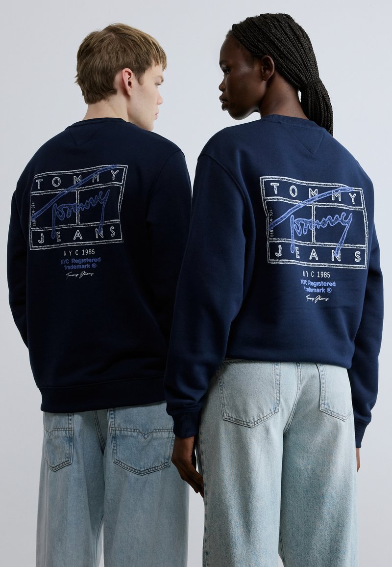 Tamnoplave majice s kapuljačom imaju bijeli i plavi tiskani logo na leđima, uparene s lagano plavim, širokim trapericama.