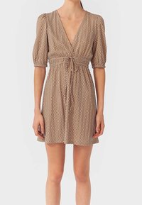 Robe courte marron à pois blancs, décolleté en V profond, manches bouffantes et taille à cordon, portée par une personne se tenant devant un fond uni.