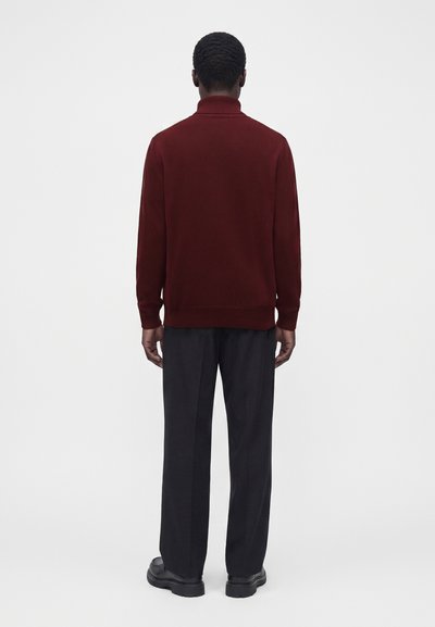 Homme vu de dos portant un pull à manches longues rouge foncé, un pantalon noir et des chaussures noires sur un fond clair uni.