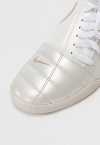 Sapatilha metálica prateada com textura acolchoada, atacadores brancos e um pequeno logo da Nike em bege na lateral. Sola de borracha com acabamento suave.