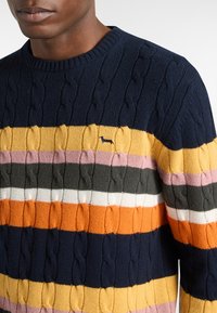 Maglione lavorato a maglia chunky con strisce orizzontali colorate in blu navy, arancione, giallo, rosa, grigio e bianco. Presenta un design a trecce texture e un piccolo logo.