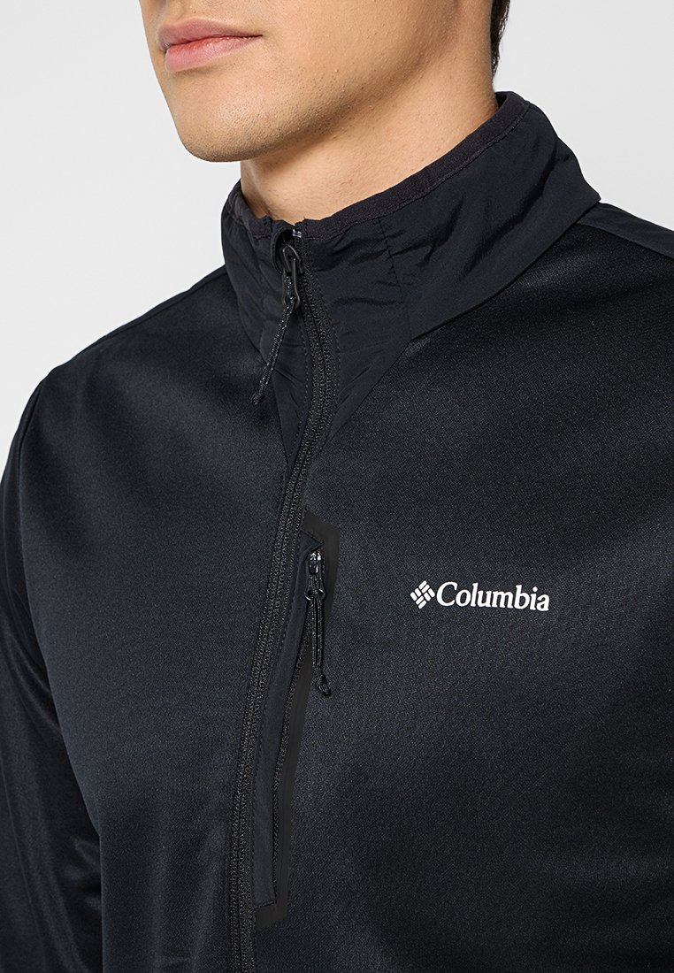 Mężczyzna w czarnej kurtce zip z wysokim kołnierzem marki Columbia, z widocznym zamkiem na kieszeni piersiowej, na białym tle.