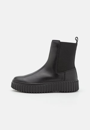 Bottine noire avec un dessus en cuir lisse, des panneaux latéraux élastiques et une semelle en caoutchouc texturé présentant des rainures horizontales.
