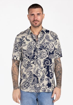 Man met kort haar en tatoeages, gekleed in een blauw overhemd met korte mouwen en een paisley-patroon, en een blauwe spijkerbroek, staand tegen een witte achtergrond.