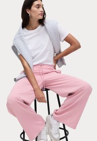 Rosa vida-byxor med dragsko i midjan, kombinerat med en vit t-shirt och en grå ärmlös tröja, buren med vita sneakers.