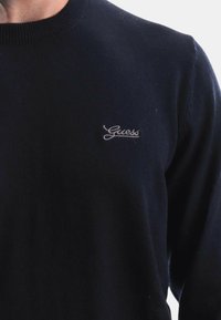 Maglione blu navy realizzato in morbido tessuto, con un colletto a coste e un piccolo logo ricamato "Guess" sul lato sinistro del petto.