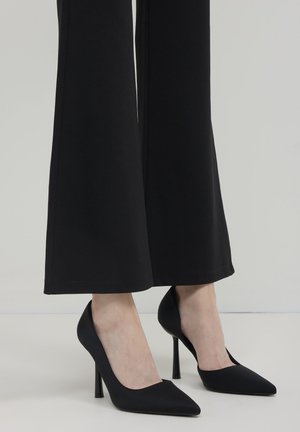 Scarpe nere con tacco alto e punta affilata, realizzate in materiale liscio, appoggiate su uno sfondo chiaro, abbinate a pantaloni neri a gamba larga.