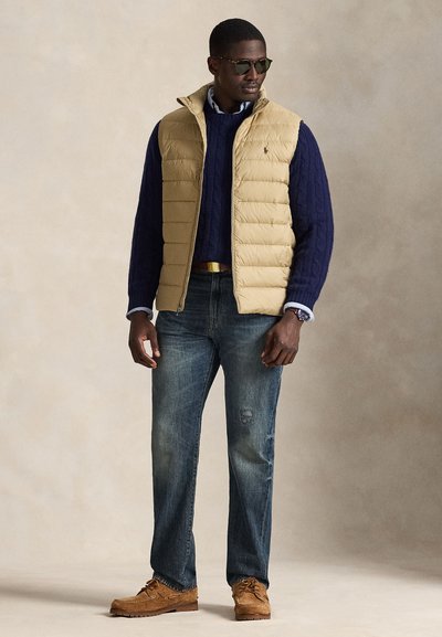 Bēša siltināta veste virs tumši zilas kabeļa mezgla džempera, saskaņota ar izbalējušām zilām džinsu biksēm un brūnām kurpēm. Aksesuāri ietver saulesbrilles un pulksteni.