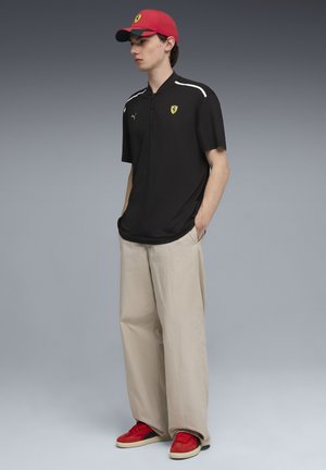 Polo noir à manches courtes, col zippé avec accents blancs ; pantalon large beige ; casquette rouge ; baskets rouges et noires avec semelles beiges.