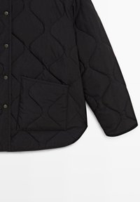 Sort quiltet jakke med trykknapper, stor frontlomme og buet kant, vist på hvid baggrund.
