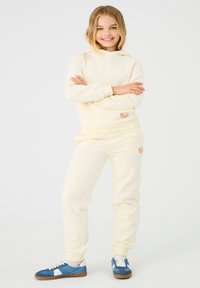 Sweat à capuche couleur crème avec pantalon de survêtement assorti. La tenue présente une texture douce et des poignets élastiques. Détails remarquables : des patchs de marque colorés.