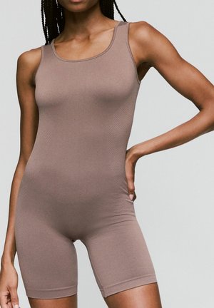 Woman wearing sleeveless, taupe-colored seamless fitted romper with shorts, standing with one hand on hip against plain background.