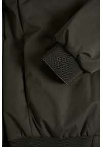 Dunkelgrüne Jacke mit strukturiertem, geripptem Bündchen. Das Material wirkt glatt und wasserabweisend, entworfen für Funktionalität und Komfort.
