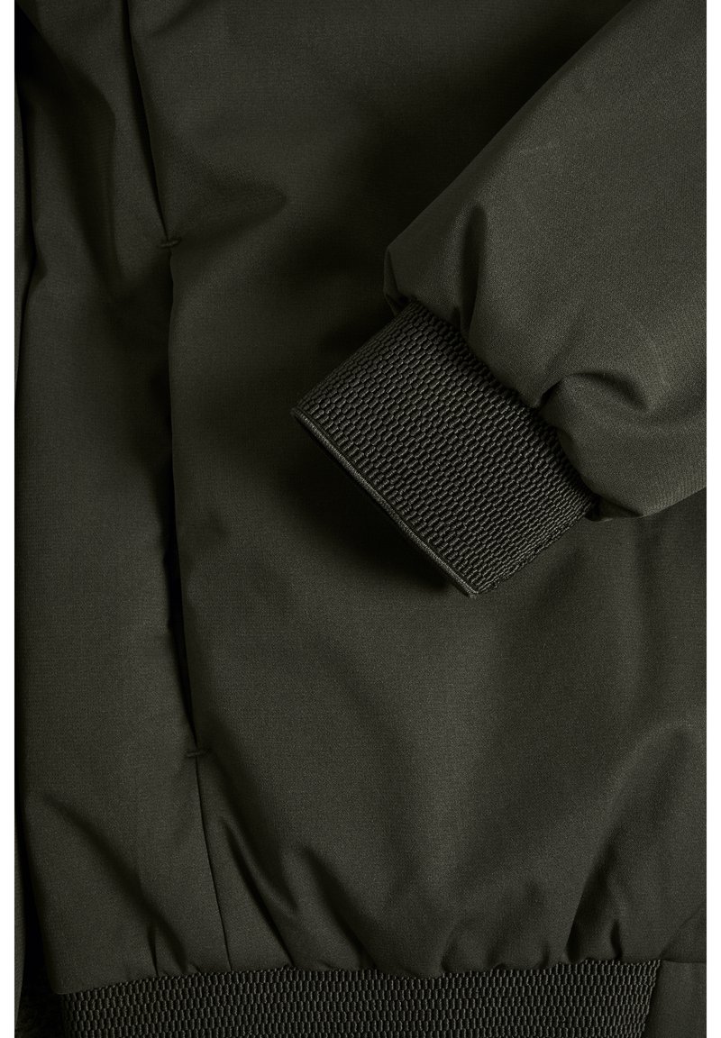 Veste vert foncé avec un poignet texturé et côtelé. Le matériau semble lisse et résistant à l'eau, conçu pour la fonctionnalité et le confort.