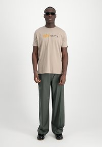 Camiseta de algodón beige con el logo "Alpha Industries" en negro y naranja, combinada con pantalones verdes holgados y zapatos negros. Lleva gafas de sol.