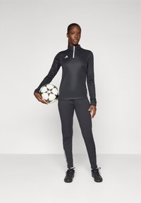 Svart atletisk topp med halv-glidelås krage og logo, sammen med matchende slim-fit bukser og svarte fotballsko, holder en mønstrete fotball.