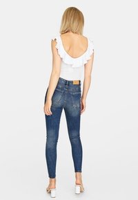 Vitt bodysuit med volangärmar, parat med blå slim-jeans. Bakifrån syns en djup u-ringning och minimala detaljer i metall. Vita klackar.