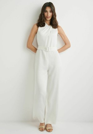 Kobieta w białym, bezrękawnym kombinezonie z paskiem i beżowych sandałach stoi przy białej ścianie, z rękami za plecami.