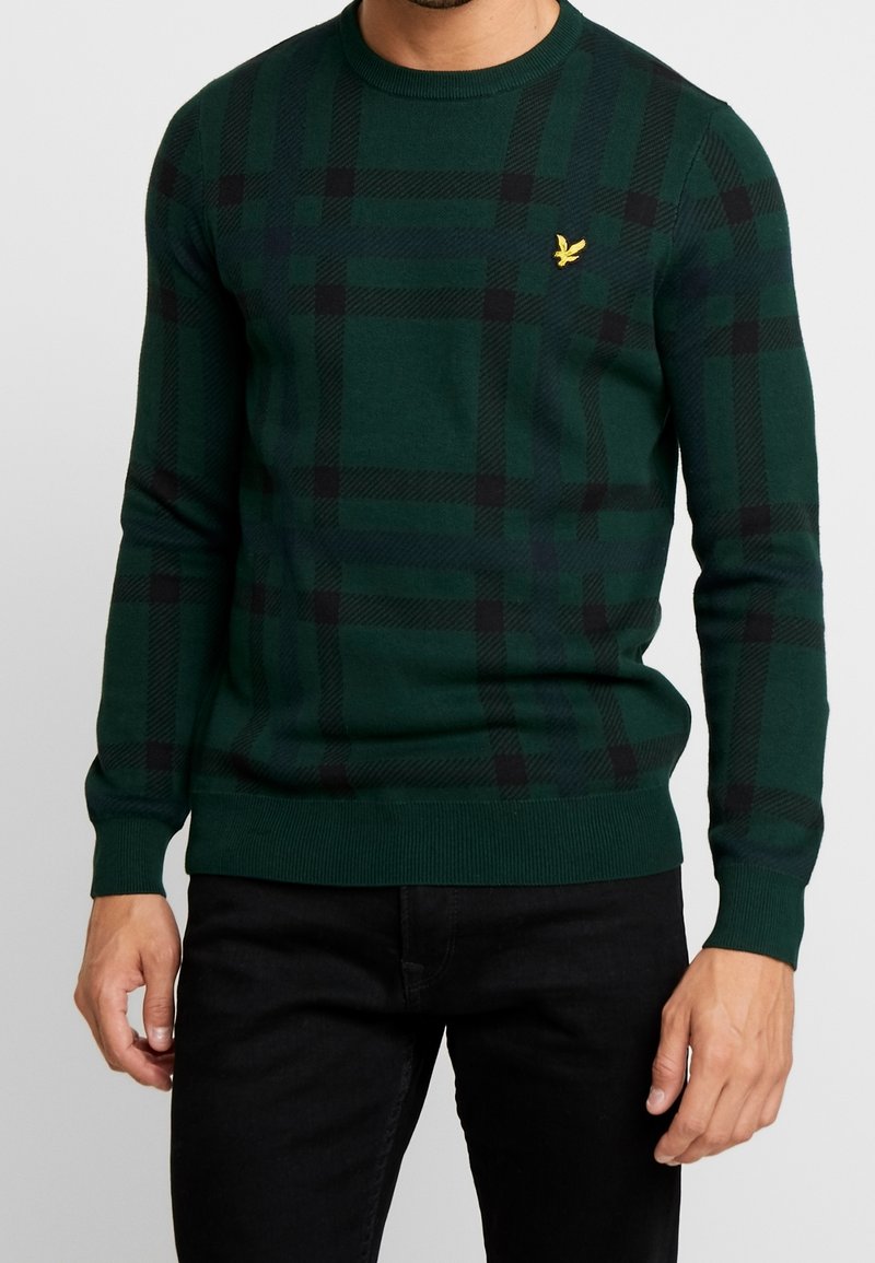 Homme portant un pull à carreaux vert foncé et noir avec un logo d’oiseau jaune sur la poitrine, associé à un pantalon noir.