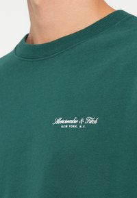 Tamnozelena pamučna majica s kratkim rukavima, s bijelim izvezenim logotipom "Abercrombie & Fitch" i natpisom "NEW YORK, N.Y." ispod. Glatka tekstura.