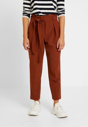 Kobieta ubrana w rude spodnie z wysokim stanem związywane paskiem, do tego czarno-biała koszula w paski z długim rękawem oraz białe trampki.